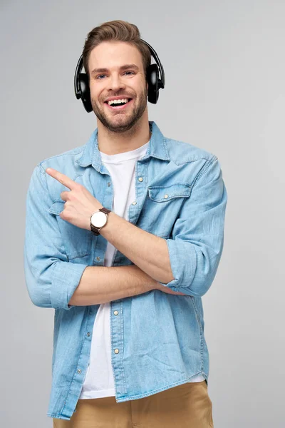 Pria tampan yang bahagia dengan kemeja jins menunjuk berdiri melawan latar belakang abu-abu mengenakan headphone besar — Stok Foto