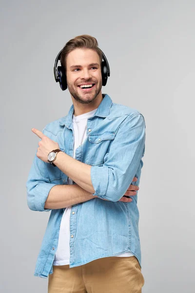 Щасливий молодий красивий чоловік у джинсовій сорочці, що вказує, що стоїть на сірому фоні в великих навушниках — стокове фото