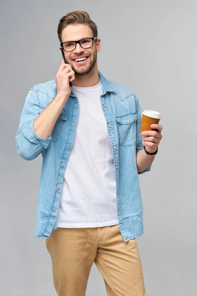 Ganska avslappnad man i blå jeans skjorta hålla sin telefon och kopp kaffe för att gå stående över studio grå bakgrund — Stockfoto