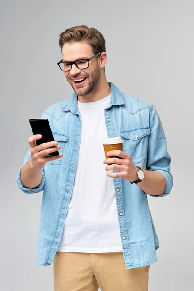 Hombre bastante casual en camisa vaqueros azules sosteniendo su teléfono y una taza de café para ir de pie sobre fondo gris estudio — Foto de Stock