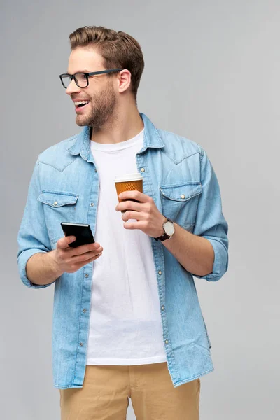 Homem muito casual em camisa jeans azul segurando seu telefone e xícara de café para ir de pé sobre estúdio fundo cinza — Fotografia de Stock
