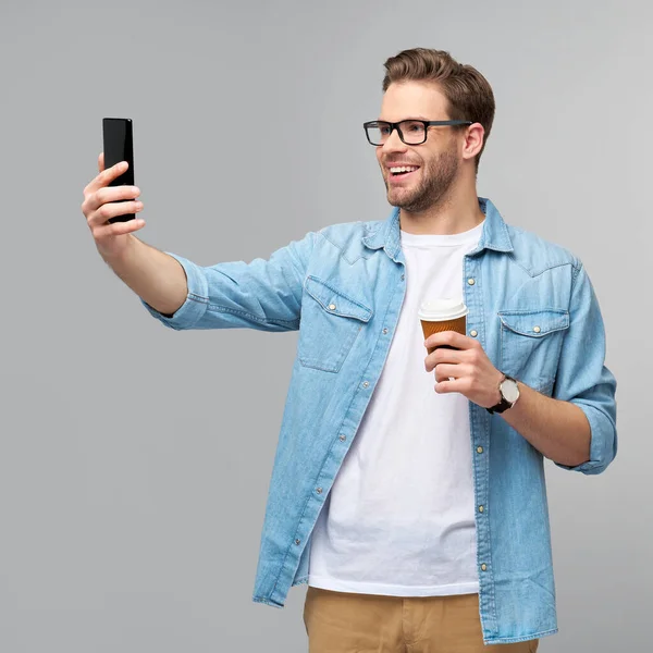 Vrij casual man in blauwe jeans shirt met zijn telefoon en kopje koffie te gaan staan over studio grijze achtergrond — Stockfoto