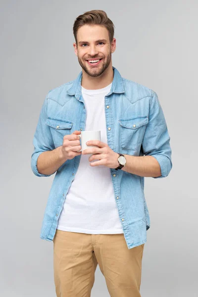 Retrato de jovem caucasiano bonito em camisa jeans sobre fundo claro segurando xícara de café — Fotografia de Stock