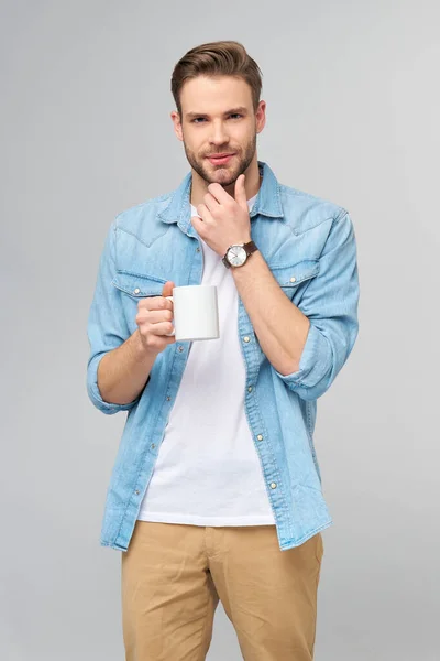 Retrato de joven guapo hombre caucásico en jeans camisa sobre fondo claro sosteniendo taza de café — Foto de Stock