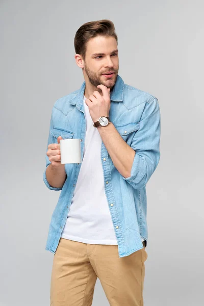Retrato de joven guapo hombre caucásico en jeans camisa sobre fondo claro sosteniendo taza de café —  Fotos de Stock
