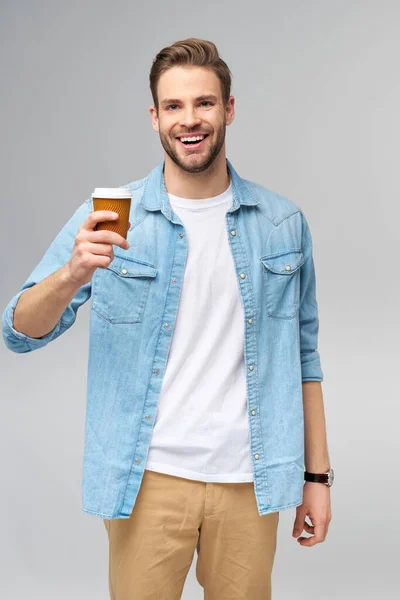 Retrato de jovem caucasiano bonito em camisa jeans sobre fundo claro segurando xícara de café para ir — Fotografia de Stock