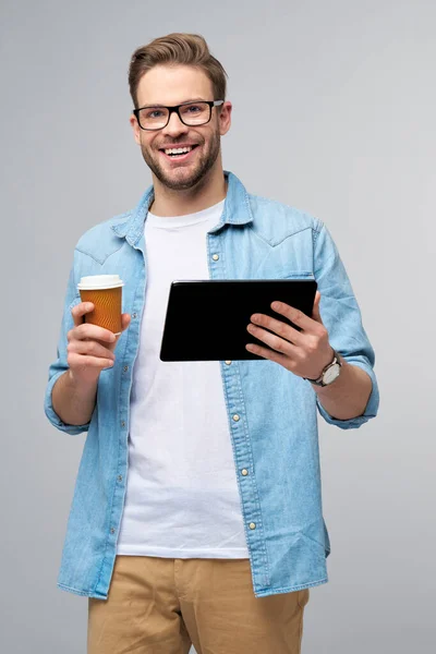Jovem feliz vestindo camisa jeans de pé e usando tablet pc pad e segurando xícara de café para ir de pé sobre estúdio fundo cinza — Fotografia de Stock