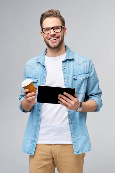 Jeune homme heureux portant une chemise en jean debout et utilisant une tablette pc pad et tenant une tasse de café pour aller debout sur fond gris studio — Photo