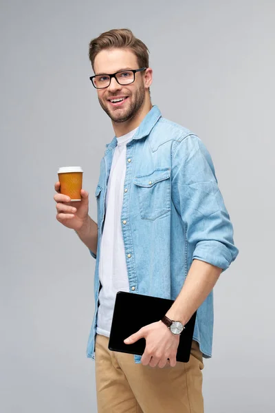 Ευτυχισμένος νεαρός άνδρας φορώντας τζιν πουκάμισο στέκεται και χρησιμοποιώντας tablet pc pad και κρατώντας φλιτζάνι καφέ για να πάει στέκεται πάνω από studio γκρι φόντο — Φωτογραφία Αρχείου