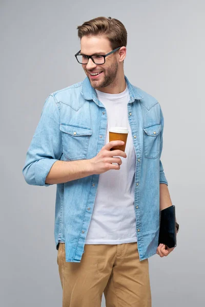 Щасливий молодий чоловік у джинсовій сорочці стоїть і використовує планшетну накладку і тримає чашку кави, щоб перейти на сірий фон студії — стокове фото