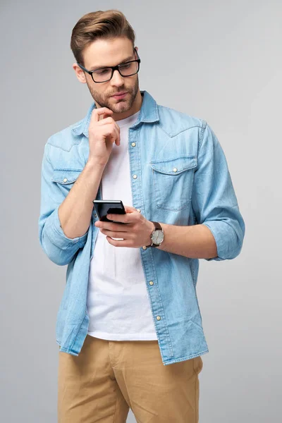 Bastante casual hombre en azul jeans camisa sosteniendo su teléfono de pie sobre estudio gris fondo — Foto de Stock