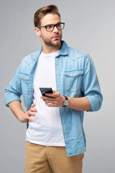 一个穿着蓝色牛仔裤衬衫的非常随意的男人，拿着手机站在工作室灰色的背景上 — 图库照片