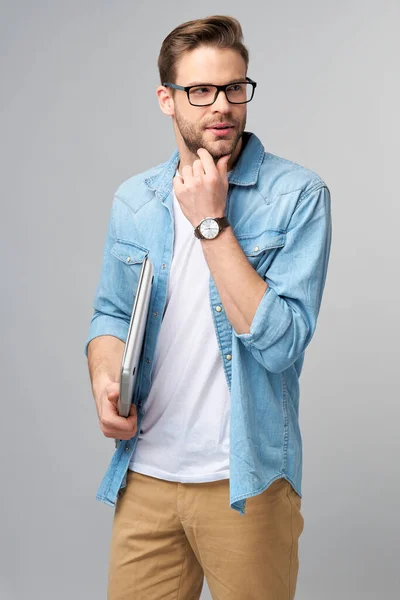 Homem barbudo jovem concentrado usando óculos vestidos com camisa jeans segurando laptop isolado sobre fundo estúdio cinza — Fotografia de Stock