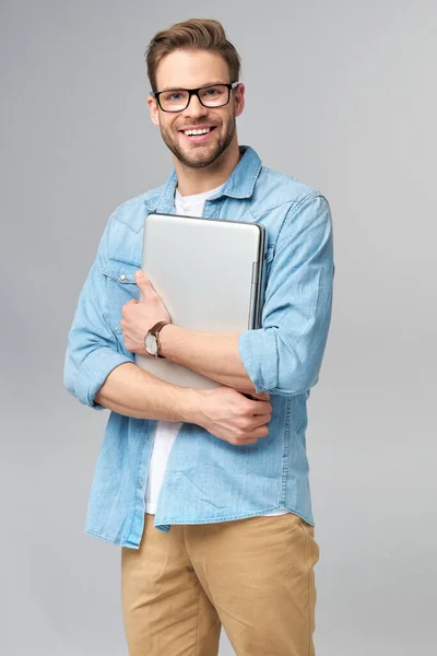 Konzentrierter junger bärtiger Mann mit Brille, Jeanshemd und Laptop isoliert vor grauem Studiohintergrund — Stockfoto
