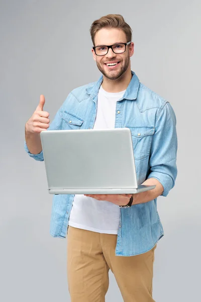 Hombre barbudo joven concentrado con gafas vestidas con camisa vaquera que sostiene el ordenador portátil aislado sobre fondo gris del estudio —  Fotos de Stock