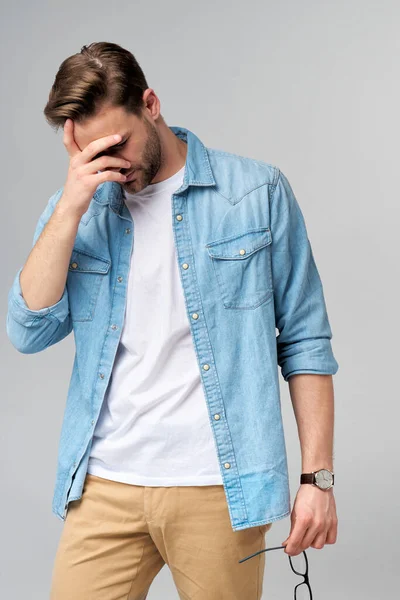 Frustrado joven caucásico hombre en jeans camisa tocando la cabeza con la mano mientras está de pie sobre fondo gris estudio —  Fotos de Stock