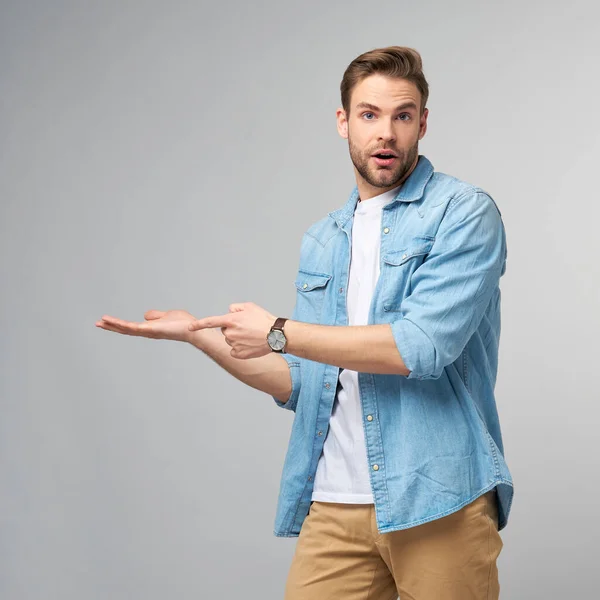 Feliz jovem bonito homem de camisa jeans apontando para longe de pé contra fundo cinza — Fotografia de Stock