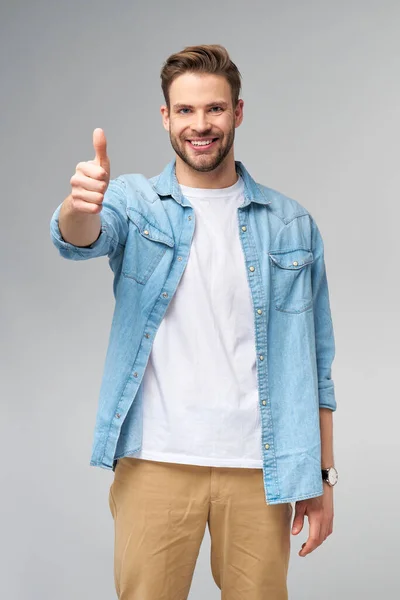 Retrato de jovem bonito caucasiano homem em jeans camisa mostrando grande polegar para cima gesto de pé sobre luz fundo — Fotografia de Stock