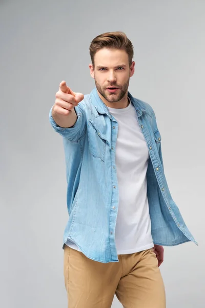 Feliz joven guapo hombre en jeans camisa apuntando a la cámara de pie sobre fondo gris — Foto de Stock