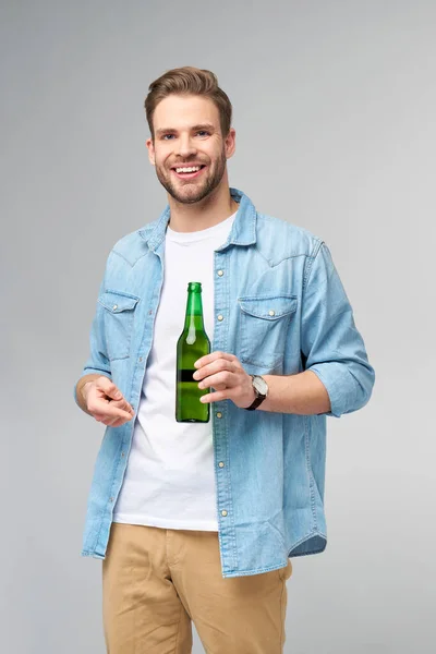 Homem jovem segurando vestindo camisa jeans segurando Garrafa de cerveja em pé sobre fundo cinza — Fotografia de Stock