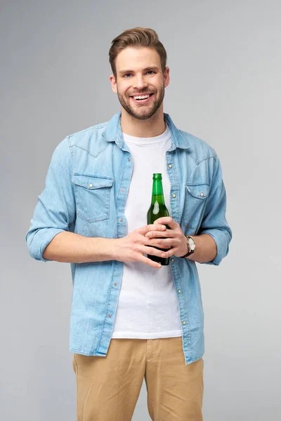 Молодий чоловік тримає в джинсах сорочку, тримаючи пляшку пива над сірим фоном. — стокове фото