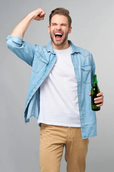 Homem jovem segurando vestindo camisa jeans segurando Garrafa de cerveja em pé sobre fundo cinza — Fotografia de Stock