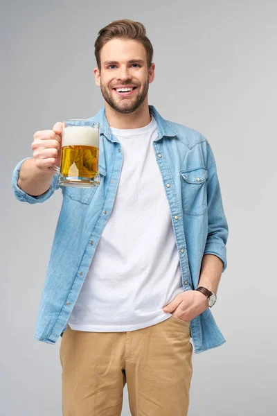 Ung man med jeans skjorta med glas öl stående över grå bakgrund — Stockfoto