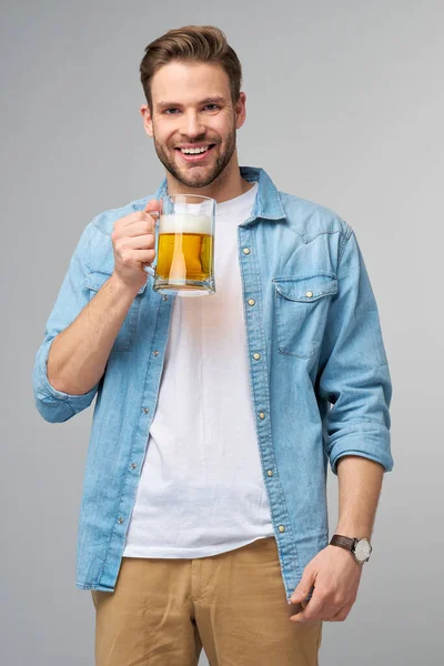 Homem jovem segurando vestindo camisa jeans segurando vidro de cerveja em pé sobre fundo cinza — Fotografia de Stock