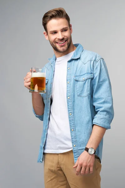 Homem jovem segurando vestindo camisa jeans segurando vidro de cerveja em pé sobre fundo cinza — Fotografia de Stock