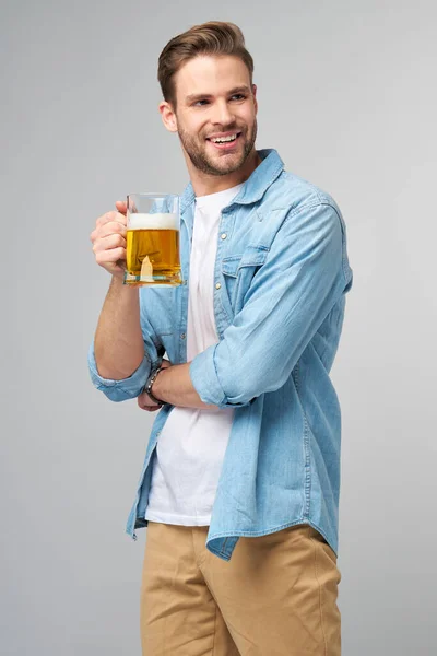 Joven con camisa vaquera sosteniendo vaso de cerveza de pie sobre fondo gris —  Fotos de Stock