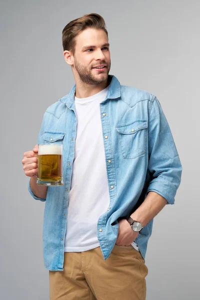 Joven con camisa vaquera sosteniendo vaso de cerveza de pie sobre fondo gris —  Fotos de Stock