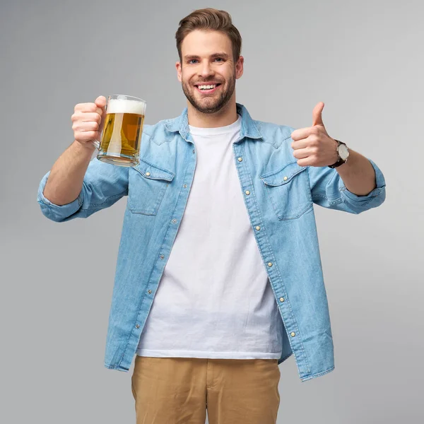 Homem jovem segurando vestindo camisa jeans segurando vidro de cerveja em pé sobre fundo cinza — Fotografia de Stock