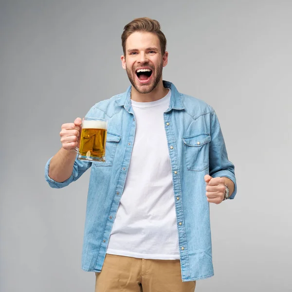 Homem jovem segurando vestindo camisa jeans segurando vidro de cerveja em pé sobre fundo cinza — Fotografia de Stock