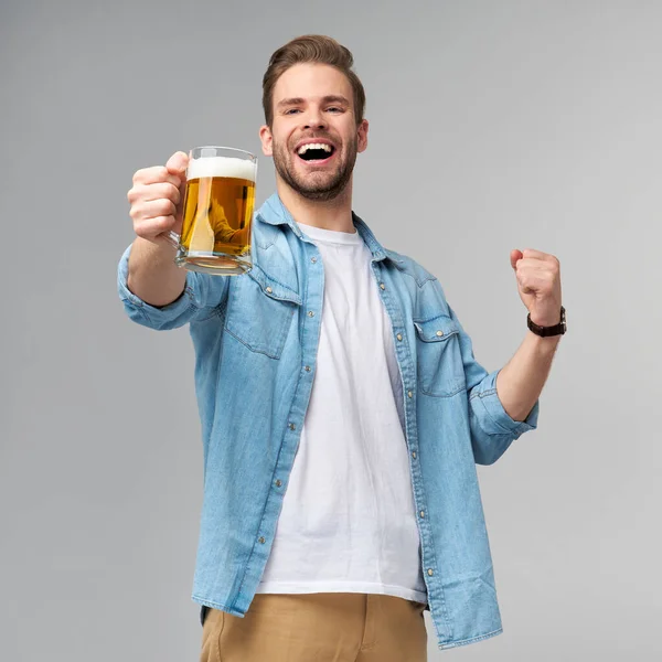 Mladý muž drží na sobě džínovou košili drží sklenici piva stojí nad šedým pozadím — Stock fotografie
