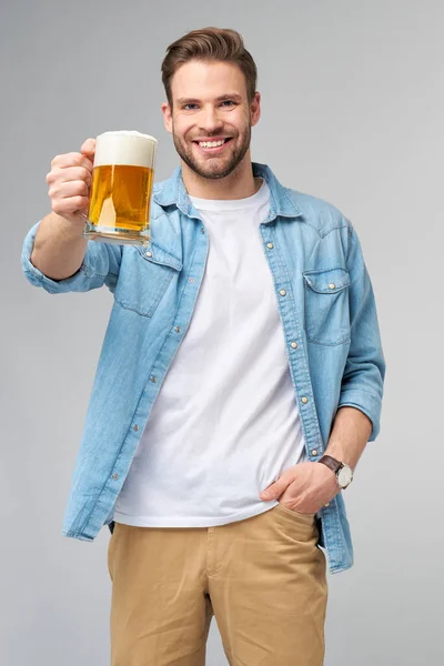 Mladý muž drží na sobě džínovou košili drží sklenici piva stojí nad šedým pozadím — Stock fotografie