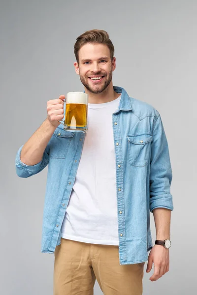 年轻男子穿着牛仔裤衬衫，举着一杯啤酒，站在灰色的背景下 — 图库照片