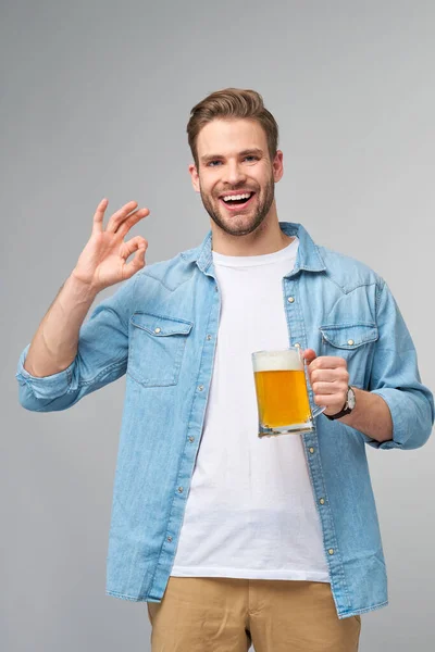 Homem jovem segurando vestindo camisa jeans segurando vidro de cerveja em pé sobre fundo cinza — Fotografia de Stock