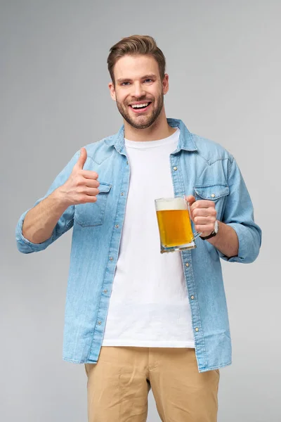 Joven con camisa vaquera sosteniendo vaso de cerveza de pie sobre fondo gris —  Fotos de Stock