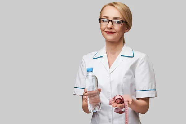 Porträt einer jungen kaukasischen Ärztin mit Wasserflasche und Maßband vor hellgrauem Hintergrund — Stockfoto