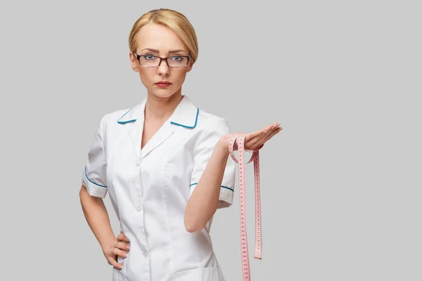 Mujer alegre médico o enfermera sosteniendo una cinta métrica de pie sobre fondo gris —  Fotos de Stock