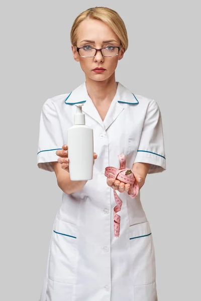 Retrato de especialista en belleza de pie sobre fondo gris y sosteniendo en su botella de mano de loción corporal y cinta métrica —  Fotos de Stock