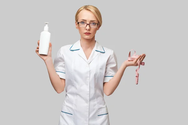 Portrait of beauty specialist standing against grey background and holding in her hand bottle of body lotion and measure tape — Stock Photo, Image