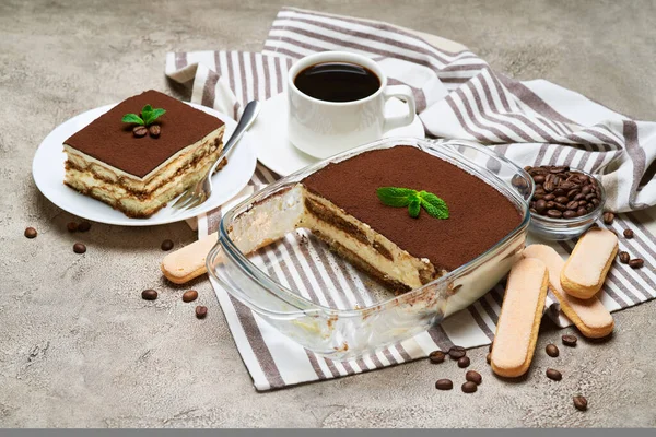 Traditional Italian Tiramisu dessert in glass baking dish, savoiardi cookies and portion on grey concrete background — Stock Photo, Image