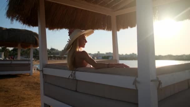 Mujer sentada en Cabana con techo de paja en una playa de arena al atardecer — Vídeo de stock