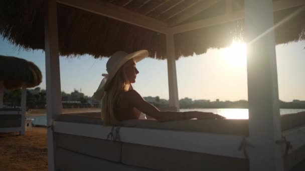 Kvinnan sitter vid Cabana med halmtak på en sandstrand vid solnedgången — Stockvideo