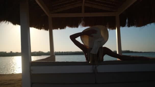 Gün batımında kumlu bir sahilde saman çatısı olan Cabana 'da oturan bir kadın. — Stok video