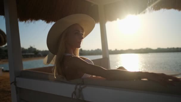 Mujer sentada en Cabana con techo de paja en una playa de arena al atardecer — Vídeo de stock