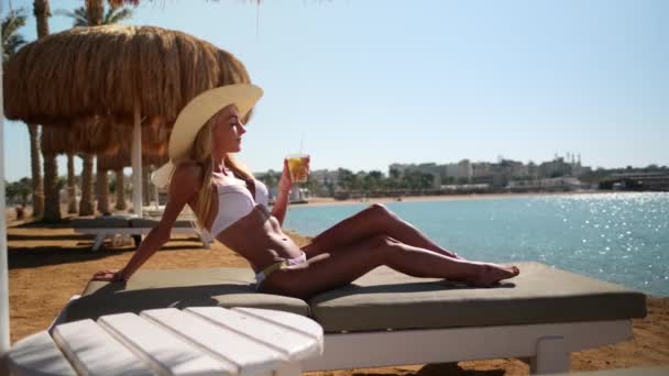 Sexy vrouw in bikini zittend op een ligstoel onder stro parasol op het strand met een glas met cocktail of drankje — Stockvideo