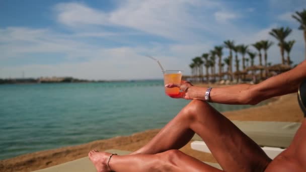 Kvinna klädd i bikini sittandes på solstol under halmtak paraply på stranden — Stockvideo