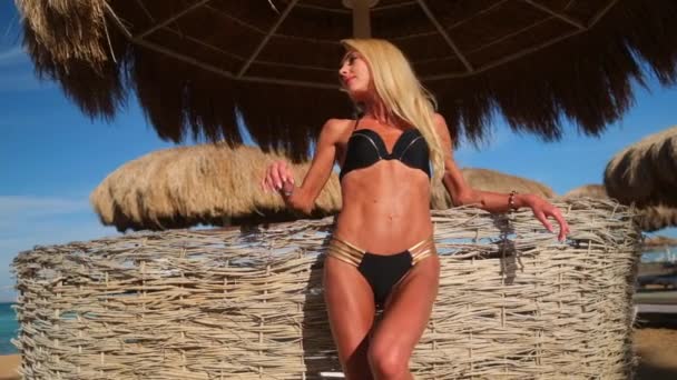 Сексуальная женщина в бикини, стоящая под соломенным навесом зонта на пляже — стоковое видео